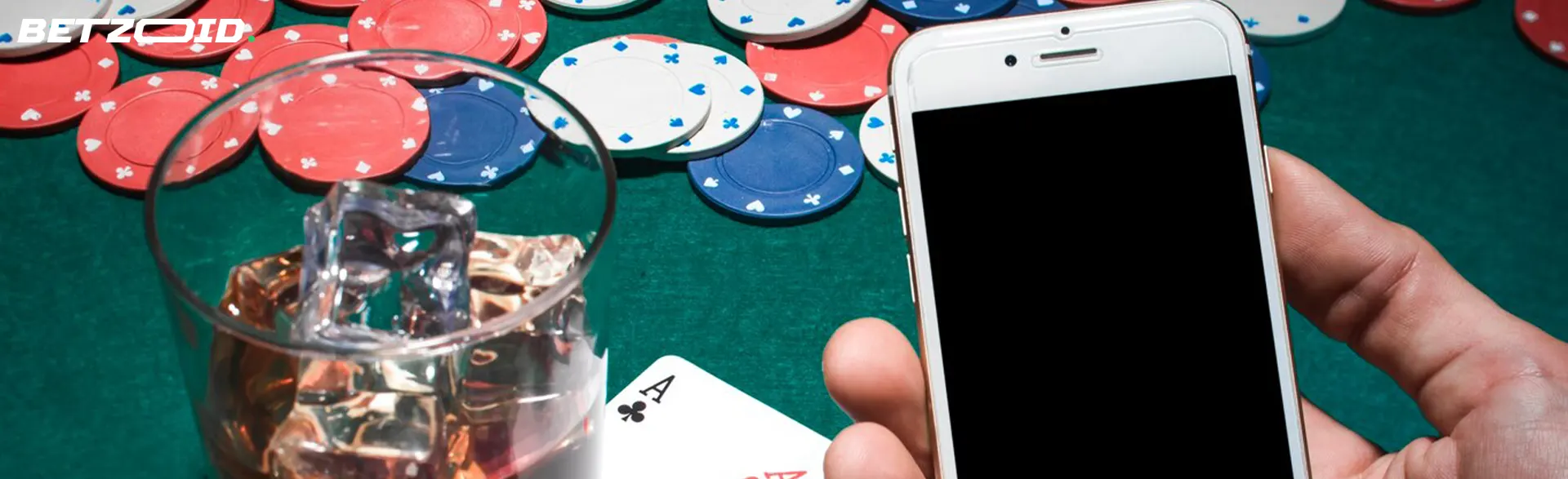 A smartphone in the hands of a player on the background of casino chips and next to a glass of drink.